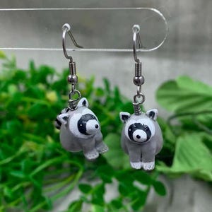 May include: A pair of dangle earrings featuring small, sculpted gray and white raccoons with black eye masks. The raccoon charms are attached to silver-toned metal fishhook wires, designed for pierced ears, showcasing detailed facial features.