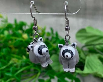 May include: A pair of dangle earrings featuring small, sculpted gray and white raccoons with black eye masks. The raccoon charms are attached to silver-toned metal fishhook wires, designed for pierced ears, showcasing detailed facial features.