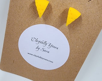 Può includere: Un paio di orecchini a bottone a forma di spicchio di formaggio giallo su una scheda marrone. Gli orecchini hanno piccoli fori, che assomigliano al formaggio. La scheda ha un adesivo ovale bianco con "Playfully Yours by Sara" e un sito web.