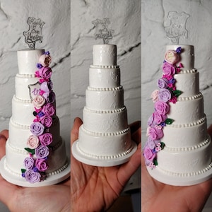 May include: A miniature five-tier white wedding cake featuring embossed floral patterns, beaded borders, and a cascading arrangement of purple, pink, and white roses. A clear acrylic cake topper displays the text Happily Ever Wooten.