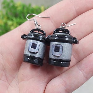 May include: A pair of dangle earrings featuring miniature replicas of a black and silver pressure cooker. Each charm has a glossy black lid and base with a textured silver body and a small digital display panel on the front, attached to silver hooks.