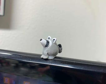 May include: A small, gray and white raccoon figurine with a black mask around its eyes, perched on the top edge of a black computer monitor. The raccoon has a rounded body, pointed ears, and a small black nose, facing upward and to the left.