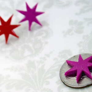 May include: Three eight-pointed star shapes, two in magenta and one in red, are displayed. One magenta star is positioned on top of a United States quarter coin for scale, while the other two stars are placed separately nearby.