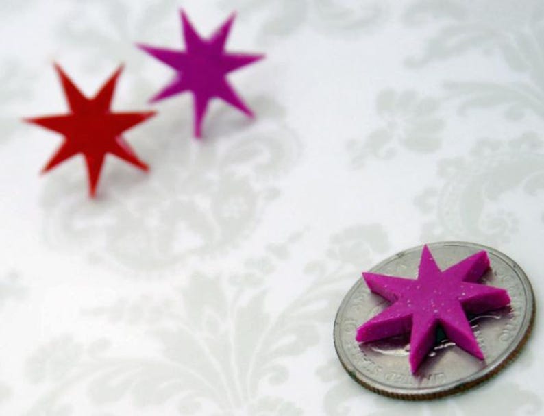 May include: Three eight-pointed star shapes, two in magenta and one in red, are displayed. One magenta star is positioned on top of a United States quarter coin for scale, while the other two stars are placed separately nearby.