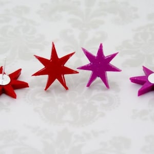 May include: Four star-shaped stud earrings arranged in a row. Two earrings are bright red and two are vibrant magenta. Each star has seven points and a smooth, glossy finish. The silver-toned metal earring posts are visible on the back of two stars.