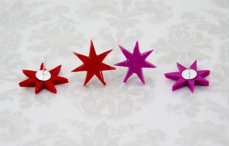 May include: Four star-shaped stud earrings arranged in a row. Two earrings are bright red and two are vibrant magenta. Each star has seven points and a smooth, glossy finish. The silver-toned metal earring posts are visible on the back of two stars.
