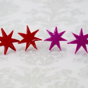 May include: Four star-shaped stud earrings arranged in a horizontal row. The two earrings on the left are a solid, glossy red color, while the two earrings on the right are a solid, glossy magenta color. Each star features eight sharp points.