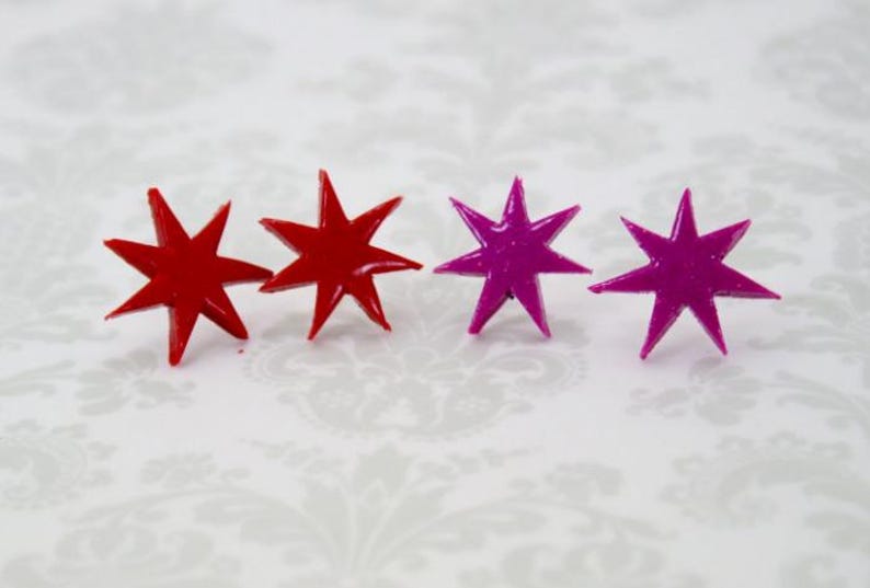 May include: Four star-shaped stud earrings arranged in a horizontal row. The two earrings on the left are a solid, glossy red color, while the two earrings on the right are a solid, glossy magenta color. Each star features eight sharp points.