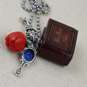 Puede incluir: Un collar de cadena plateada con un dije de manzana roja, un dije de llave plateada, un dije de espejo azul y una pequeña caja marrón con las palabras "Érase una vez" impresas en ella.