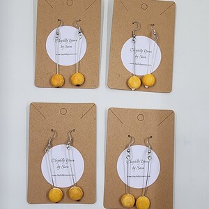 May include: Four pairs of dangle earrings with yellow and orange round ceramic beads on silver wire. The earrings are displayed on brown card stock with the text "Playfully Yours by Sara" and a website address.