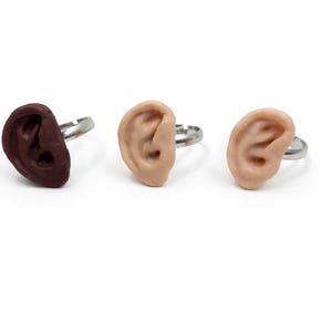 May include: Three rings with ear-shaped designs. One ring features a dark brown ear, while the other two rings have flesh-toned ears. The rings have silver-colored adjustable bands. The background is white.