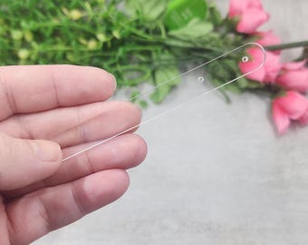 May include: A clear, rectangular acrylic nail swatch with two small holes near the rounded end. The swatch is held in a hand, with a blurred background of pink flowers and green foliage.
