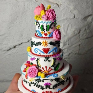 Peut inclure: Un gâteau blanc à trois niveaux avec des motifs floraux et décoratifs colorés. Le gâteau est orné de roses roses, d'accents jaunes et de motifs complexes rouges, bleus et verts. Le gâteau est tenu dans une main.