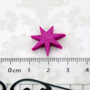 May include: A magenta six-pointed star embellishment with a subtle glitter finish. The star is positioned above a ruler, showing it measures approximately 0.75 inches in width and height from tip to tip, featuring sharp, defined points.