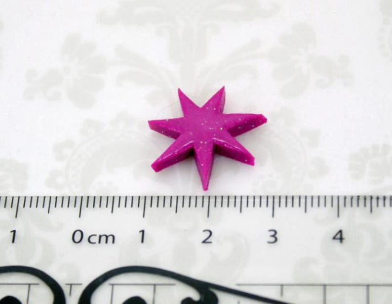 May include: A magenta six-pointed star embellishment with a subtle glitter finish. The star is positioned above a ruler, showing it measures approximately 0.75 inches in width and height from tip to tip, featuring sharp, defined points.