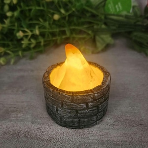 May include: A small, battery-operated LED candle with a flickering flame effect. The candle is encased in a gray, brick-patterned holder, creating a rustic appearance. The flame is a warm orange color, providing a soft glow.
