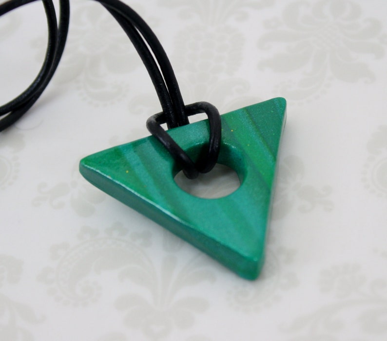 May include: A green triangle pendant with a hole in the center, hanging from a black leather cord. The pendant has a marbled effect and is made of polymer clay.