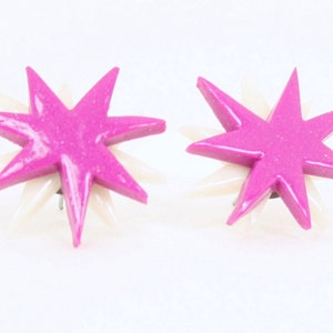Pu&ograve; includere: Un paio di orecchini a forma di stella rosa e bianca. Le stelle hanno una finitura scintillante e sono posizionate su uno sfondo bianco.