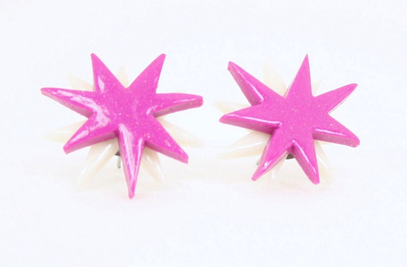 Pu&ograve; includere: Un paio di orecchini a forma di stella rosa e bianca. Le stelle hanno una finitura scintillante e sono posizionate su uno sfondo bianco.