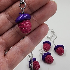 May include: A set of raspberry-themed jewellery featuring a necklace and earrings. The necklace has a silver chain and a pendant shaped like a raspberry with a purple top. The earrings are silver hoops with a raspberry charm hanging from each hoop.