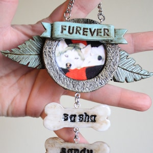 May include: A decorative hanging ornament featuring a circular frame with a photo of two dogs, flanked by two leaf-shaped accents. A light blue banner above reads FUREVER. Two bone-shaped charms hang below, inscribed with the names sasha and sandy.