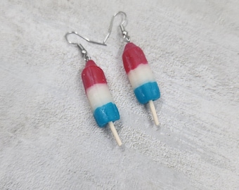 May include: A pair of dangle earrings shaped like rocket popsicles. Each earring features a red, white, and blue layered popsicle design with a small wooden stick at the bottom, suspended from silver-toned metal fishhook ear wires.