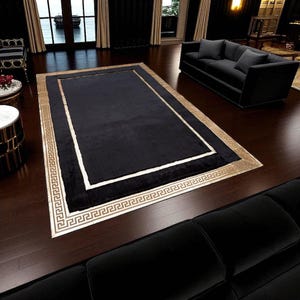 May include: A rectangular black rug with a gold border and a white inner border. The gold border features a Greek key pattern. The rug is placed on a dark wood floor in a living room setting.
