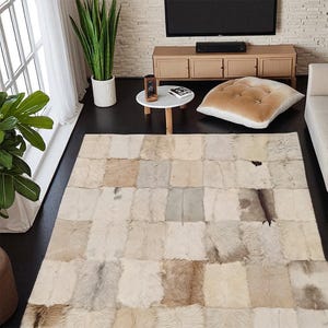 Puede incluir: Una alfombra rectangular compuesta por varios parches rectangulares en tonos crema, beige y marrón. La alfombra está colocada sobre un suelo oscuro en un salón. El diseño de la alfombra añade un elemento natural y texturizado a la decoración de la habitación.
