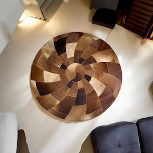 Puede incluir: Una alfombra redonda de cuero marrón con un patrón de espiral de diferentes tonos de marrón. La alfombra está hecha de piel de vaca y tiene un diseño geométrico único.