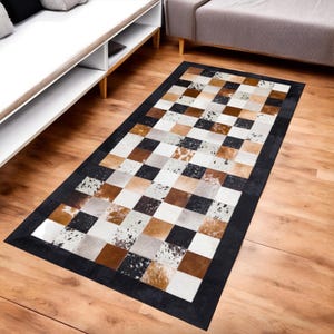 Op de afbeelding: Een rechthoekig vloerkleed met een patchwork-ontwerp. Het kleed heeft vierkanten in verschillende kleuren en texturen, waaronder bruin, zwart, wit en beige. Het heeft een zwarte rand en ligt op een houten vloer.