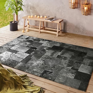 May include: A rectangular area rug with a patchwork design in shades of gray and black. The rug is made of various textures and patterns, creating a modern and textured look. The rug is placed on a wooden deck.