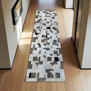 May include: A rectangular hallway rug with a geometric pattern. The rug features a patchwork design of white, grey, and black squares. The rug is placed on a light-coloured wooden floor, creating a contrast.
