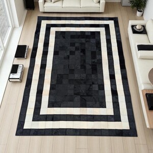 May include: A large rectangular rug with a patchwork design. The rug features a central area of black squares, framed by a white and black border. The rug is placed on a light-coloured wooden floor, with furniture visible in the background.