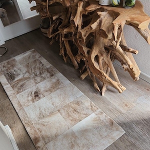 Könnte beinhalten: Ein rechteckiger Teppich mit einem Patchwork-Design aus braunen und cremefarbenen Kuhfellquadraten. Der Teppich liegt auf einem Holzboden, im Hintergrund ein großer Konsolentisch aus Naturholz. Dekorative Gegenstände befinden sich auf dem Tisch.