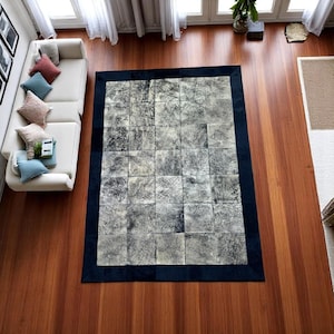 Puede incluir: Una alfombra rectangular grande con un diseño de retazos. La alfombra presenta un borde azul oscuro y un patrón central de parches cuadrados en tonos de gris y blanco. La alfombra está colocada sobre un suelo de madera.