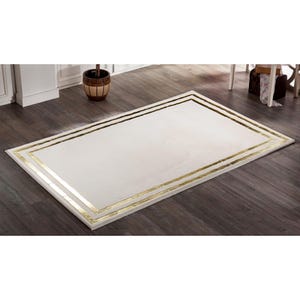 Puede incluir: Alfombra rectangular de color crema con un borde dorado doble. Se encuentra sobre un suelo de madera oscura. La alfombra mide aproximadamente 152 cm por 228 cm, adecuada para salones o dormitorios. Diseño sencillo y elegante.