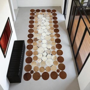 Puede incluir: Una alfombra rectangular compuesta por numerosos círculos en tonos marrones, beige y blancos. El diseño de la alfombra crea un efecto degradado, pasando del marrón oscuro al blanco en el centro. Esta alfombra decorativa es adecuada para un pasillo o entrada.