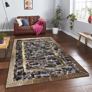 以下が含まれることがあります： テクスチャ加工された濃い茶色と黒の模様の長方形のラグ。 ラグには金の縁取りと、端にギリシャキーのデザインがあります。 ラグはリビングルームの木製の床に置かれています。