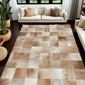 Peut inclure: Un tapis rectangulaire composé d'un assemblage de carreaux de peau de vache carrés dans des tons variés de beige, crème, brun clair et fauve. Les carreaux sont disposés en damier, offrant une surface aux nuances naturelles et texturées.