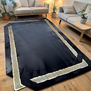May include: A rectangular black rug with a gold Greek key border. The rug is placed on a hardwood floor in a living room setting, with two sofas and plants visible in the background. The rug's design adds a touch of elegance.