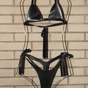 May include: A black bikini set displayed on a gold-colored wireframe mannequin. The bikini top is a halter style, and the bottoms are a thong style. The set is made of a shiny, dark material, and is hanging against a brick wall.