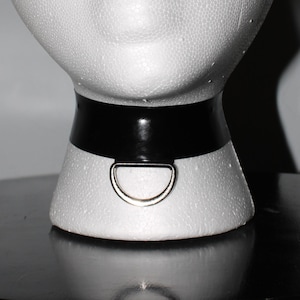 May include: A black choker collar with a silver-colored D-ring. The collar is made of a shiny, smooth material and is attached to a white foam mannequin head. The D-ring is centered on the front of the collar.
