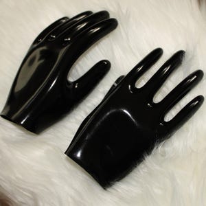 May include: Two black, glossy gloves with a hand-like shape. The gloves are made of a shiny material and are displayed on a white, fluffy surface. The gloves have a smooth, reflective surface.
