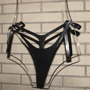 May include: Black lingerie with a unique design, suspended by a gold-colored metal frame. The garment features multiple straps and is adorned with black ribbon accents. The background is a light-colored brick wall.