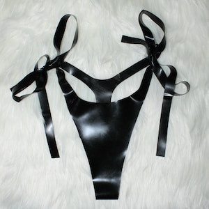 May include: Black latex lingerie set with a thong-style bottom and a halter top design. The top features straps that tie into bows at the shoulders. The shiny material contrasts with the soft, white background.
