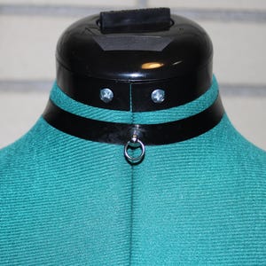 May include: A black leather choker with a silver ring closure. The choker is on a mannequin with a teal fabric covering.