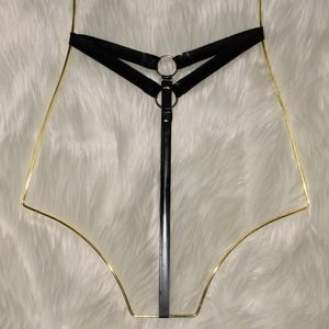 May include: Black lingerie harness with gold-colored wire frame. The harness features a black strap design with metal rings and a vertical strap. The background is a white, fluffy surface.
