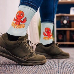 Könnte beinhalten: Ein Paar blaue und weiße Socken mit einem Lebkuchenmann-Design. Der Lebkuchenmann ist rot und orange und hat die Worte "Bite This" auf der Socke. Die Socken werden von einer Person getragen, die dunkelgrüne Sneaker trägt.