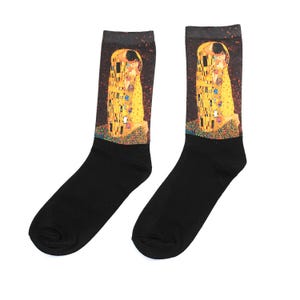 Op de afbeelding: Een paar zwarte sokken met een afdruk van Gustav Klimt's "De Kus". Het kunstwerk, met zijn goud-, geel- en bruintinten, is prominent aanwezig op het bovenste deel van de sokken.