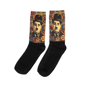 Pode incluir: Par de meias pretas com um design colorido de Charlie Chaplin, com as palavras "WHAT ART" e uma carinha triste. O fundo inclui texto em estilo grafite e um logotipo.